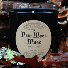 Load image into Gallery viewer, The Emerald Hearth New Moon Muse candle in black surrounded by leaves.  This candle has notes of ozone and moss.