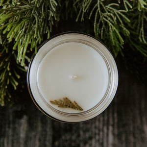 The top of the mini Flourishing Forest candle. The top of the candle has a spruce tip on it. The candle is placed next to some spruce.