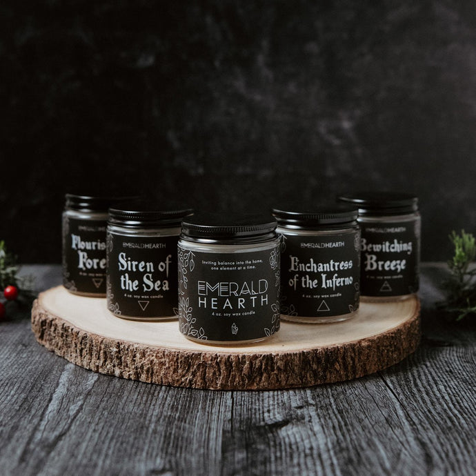 From left to right, the flourishing forest candle, siren of the sea candle, emerald hearth candle, enchantress of the inferno candle, and bewitching breeze candle displayed on a wood slat with a back background.