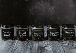 The Elemental Mini Bundle Set which includes (from left to right) the Flourishing Forest candle, Siren of the Sea candle, Emerald Hearth candle, Enchantress of the Inferno candle, and Bewitching Breeze candle all displayed on wood with a black background.