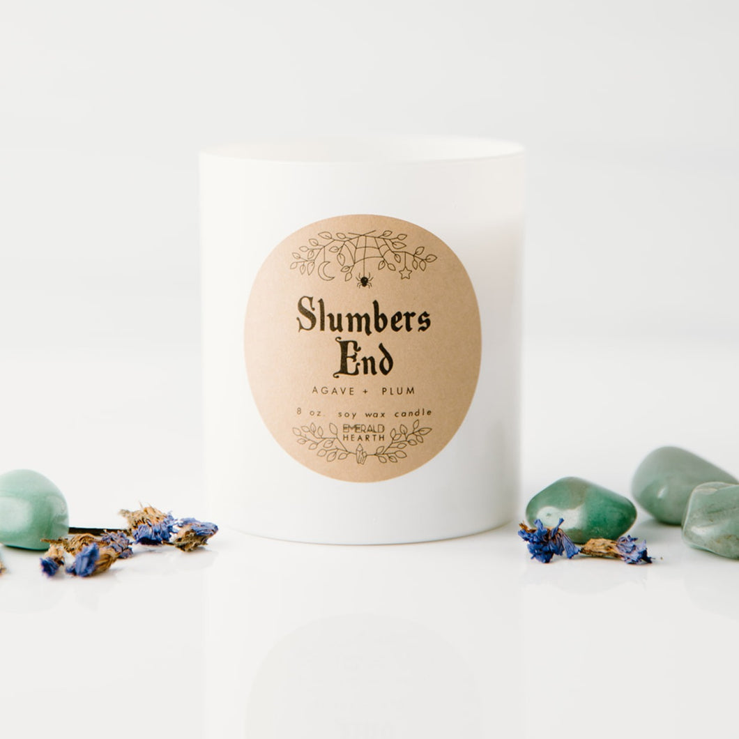 Front image of the Slumbers End candle by Emerald Hearth.  This white candle is photographed against a white background and is surrounded by flower buds and green stones.  Slumbers End is adorned with Green Aventurine and a violet Forget Me Not flower to support budding confidence in your new intentions for the coming months. 8oz 50+ hours of burn time Limited Edition Annual Release