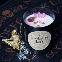 Cargar imagen en el visor de la galería, The black Everlasting Love candle by Emerald Hearth Creations. The top of the candle is showing which has rosebuds and pink quartz. The candle is on top of a Ouija board and has a gold skeleton next to it.