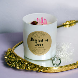 The white Everlasting Love candle with rosebuds around it photographed on top of a mirror with a ring nearby.  This candle is by Emerald Hearth Creations.
