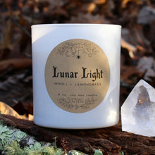 Load image into Gallery viewer, The Emerald Hearth Lunar Light candle in white on a foresty background with a crystal next to it.