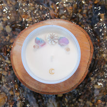 Load image into Gallery viewer, Aerial view of the Siren of the Sea candle by Emerald Hearth.  This candle is adorned with titanium-coated, pastel quartz nestled around a seashell to represent the water element.  The candle is displayed on a piece of wood on top of some water.