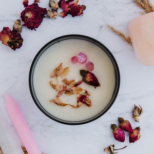 The top of the Everlasting Love candle by Emerald Hearth. The candle has rosebuds and pink quartz on top. The candle is atop a white background which has more rosebuds around it.