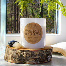 Load image into Gallery viewer, The Emerald Hearth original candle in white sitting atop a piece of wood with a window in the background.