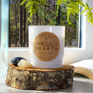 The Emerald Hearth original candle in white sitting atop a piece of wood with a window in the background.