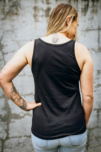 A woman wearing an Emerald Hearth tank top that says Magic Surrounds Me. The woman is standing facing away from the camera and the back of the tank top is blank.