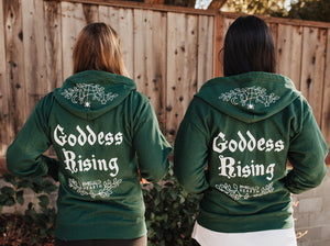 Two people wearing green Emerald Hearth hoodies facing away from the camera.  The hoodies have a spider web detail, the Emerald Hearth Logo, and the words: Goddess Rising.