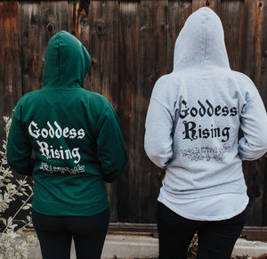 Two people wearing Emerald Hearth hoodies (one in green and one in gray) facing away from the camera.  The hoodies have a spider web detail, the Emerald Hearth Logo, and the words: Goddess Rising.