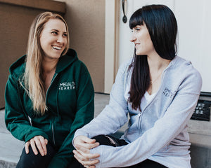 Two women wearing Emerald Hearth hoodies (one in green and one in grey) sitting down and smiling.
