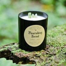 Laden Sie das Bild in den Galerie-Viewer, The black Flourishing Forest candle by Emerald Hearth creationson top of a mossy log.  The background is green. There is green quartz sticking out from the candle.