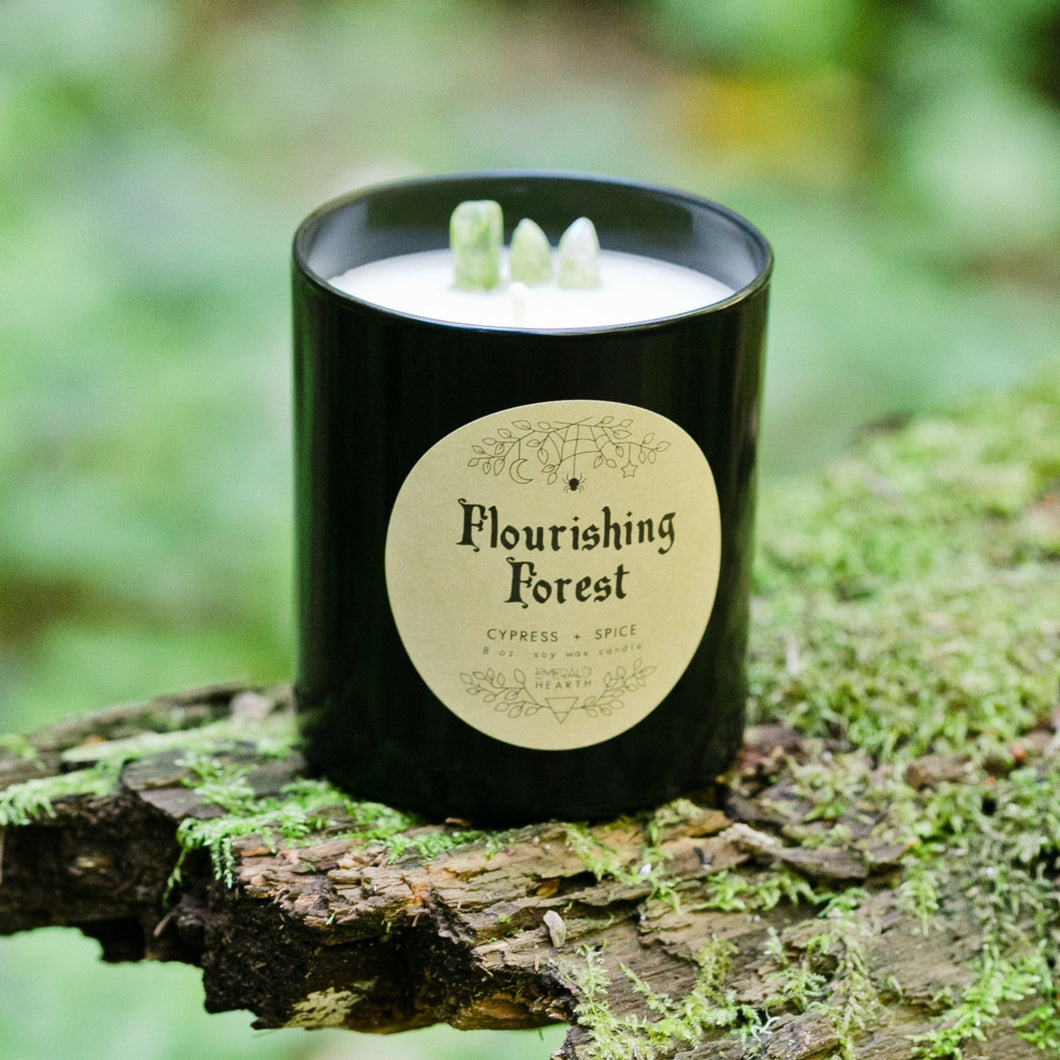 The black Flourishing Forest candle by Emerald Hearth creationson top of a mossy log.  The background is green. There is green quartz sticking out from the candle.