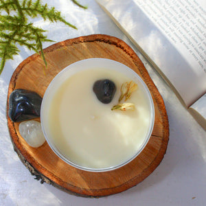 The top of the Emerald Hearth original candle topped with black onyx and jasmine buds. The candle is sitting on a slab of wood with an open book nearby.