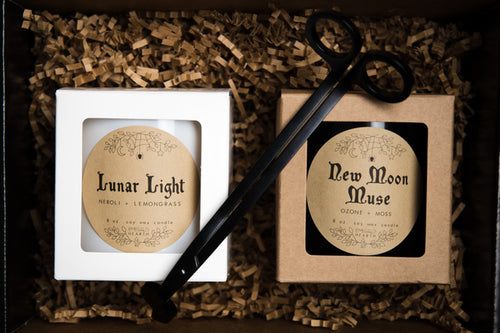 Gift packaged Lunar Love Bundle by Emerald Hearth creations.  The image shows what is included in the bundle.  The Lunar Light candle in white is on the left and the New Moon Muse candle in black is on the right.  On top of the candle is a Emerald Hearth Creations wick trimmer.