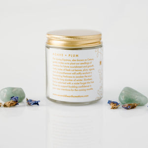 The mini version of the Slumbers End candle by Emerald Hearth Creations.  This ritual equinox candle is photographed against a white background and surrounded by Green Aventurine and violet Forget Me Not flowers.  The candle's packaging is white with gold accents.  This is the back view which tells more about this agave and plum candle.