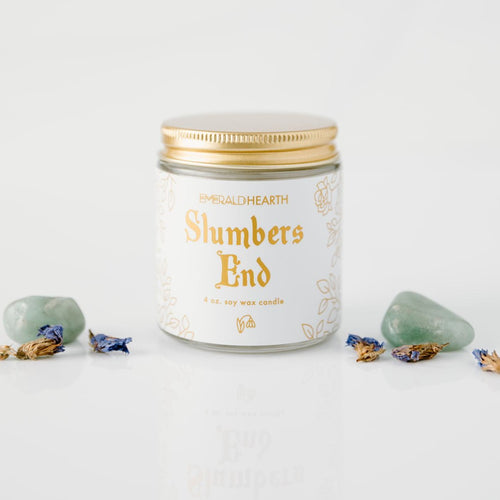 The mini version of the Slumbers End candle by Emerald Hearth Creations.  This ritual equinox candle is photographed against a white background and surrounded by Green Aventurine and violet Forget Me Not flowers.  The candle's packaging is white with gold accents.